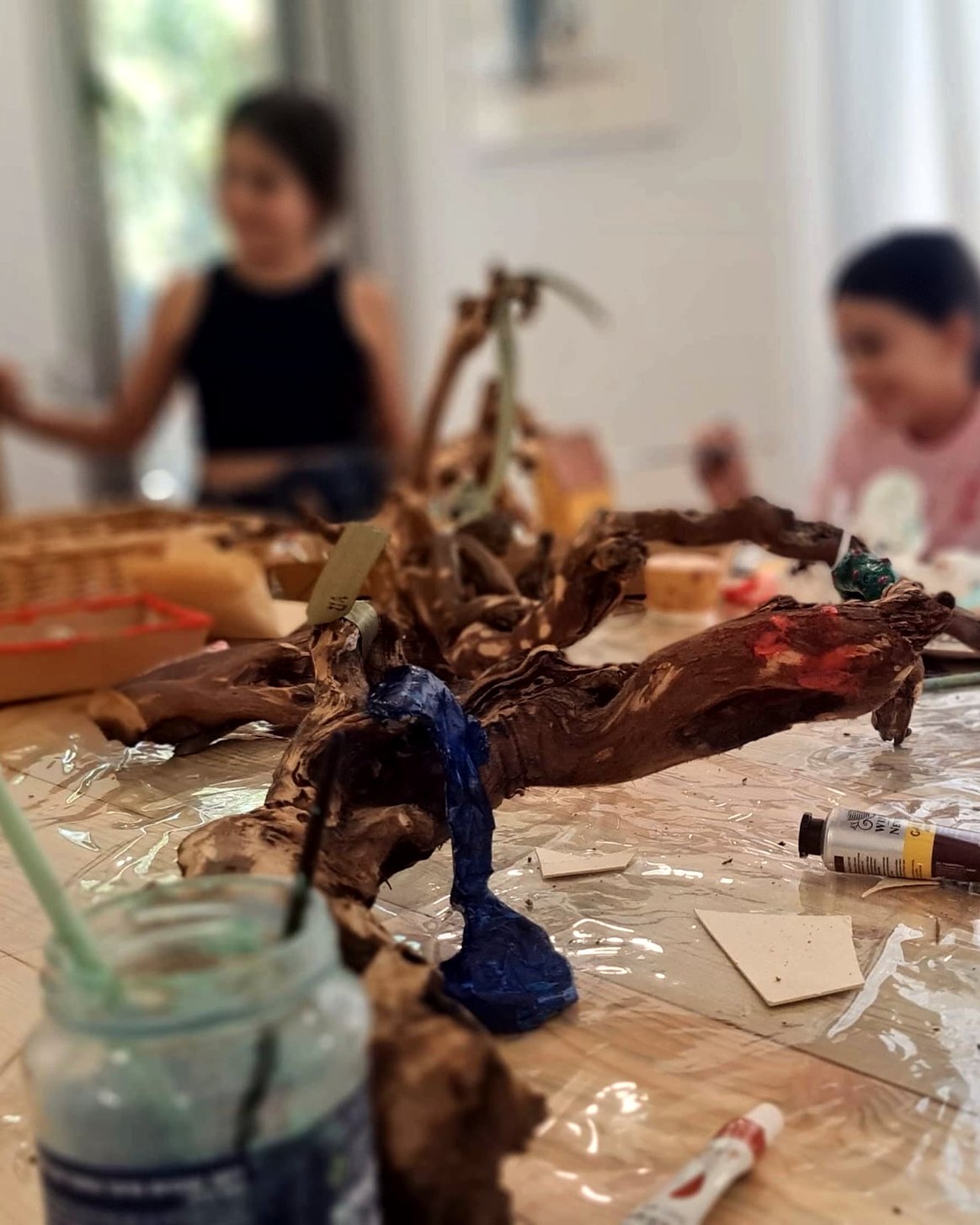 שולחן העבודה בסטודיו: ענף עץ מעוצב וחומרי יצירה, וילדות עובדות ברקע